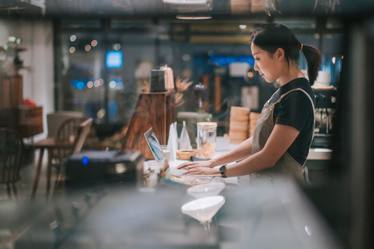 A woman in a restaurant behind the front counter on a laptop.