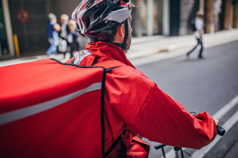 A person with red jacket on a bike delivering a restaurant order. 