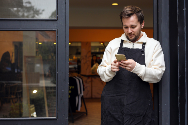 A man at the front door of a restaurant looking at his phone. 